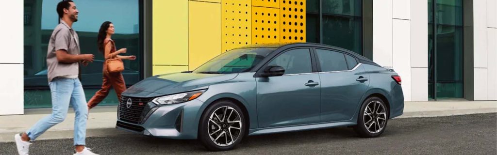 Gray sedan parked curbside in front of a modern building, with two pedestrians walking past.